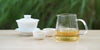 From left to right, a gaiwan, two teacups, and a glass server. The teacups and server have white tea in them.