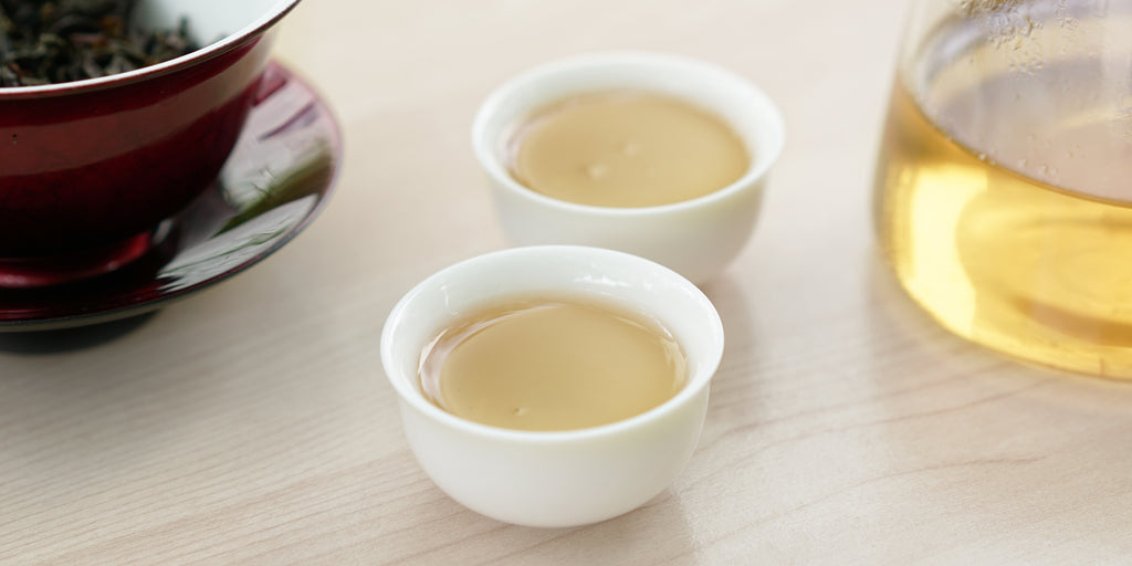 Two small teacups of Phoenix oolong tea, with a gaiwan in the background on the left and a glass server on the right.